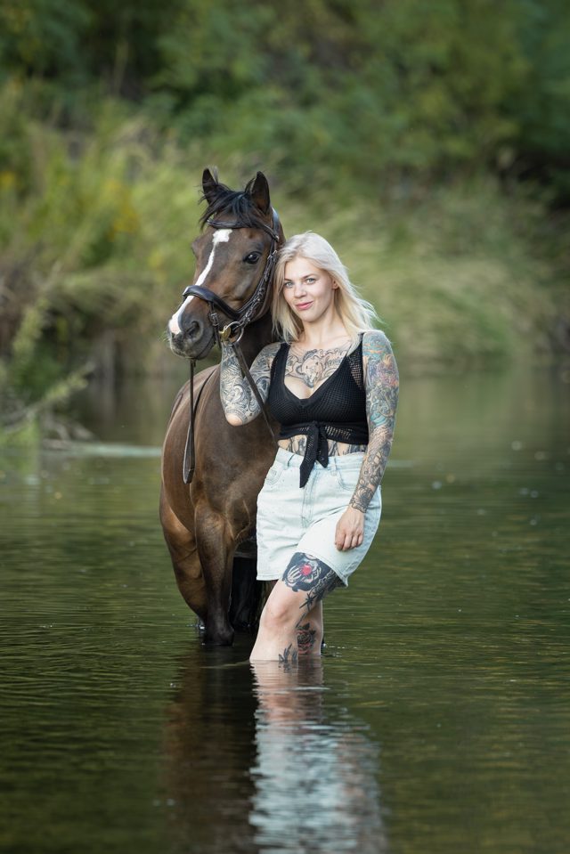Junge Frau mit blonden Haaren und Tattoos steht mit ihrer braunen Stute im Fluss, grüne Bäume im Hintergrund