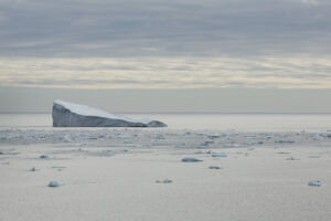 Ein Eisberg auf der linken Seite des Bildes, rundherum schwimmen kleine Einsstücke im Wasser