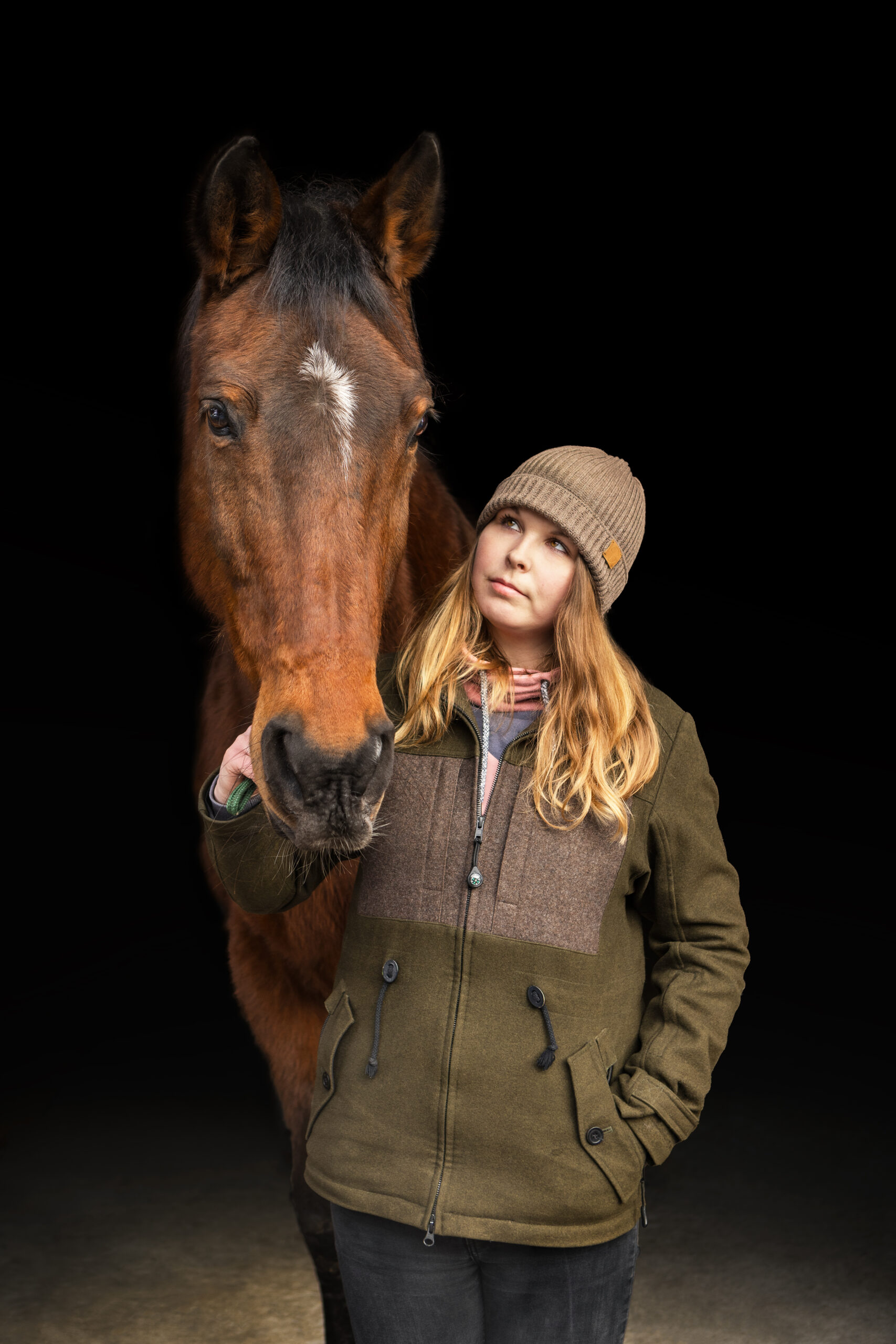 Eine braune Stute und ihre Besitzerin stehen vor einem schwarzen Hintergrund. Die Junge Frau sieht die Stute direkt an