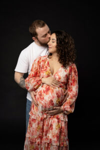 Junge schwangere Frau in buntem Kleid wird von ihrem Mann umarmt. Beide stehen vor schwarzem Hintergrund