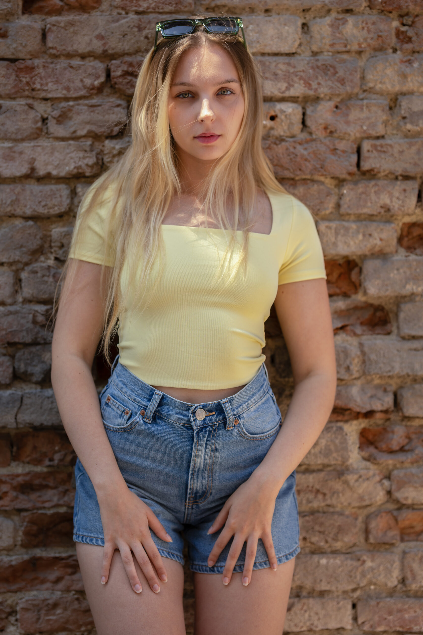 Junge blonde Frau in gelbem Shirt steht vor einer Ziegelwand und schaut in die Kamera