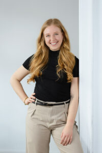 Junge rothaarige Frau mit schwarzem Shirt steht angelehnt an die Wand und lächelt in die Kamera