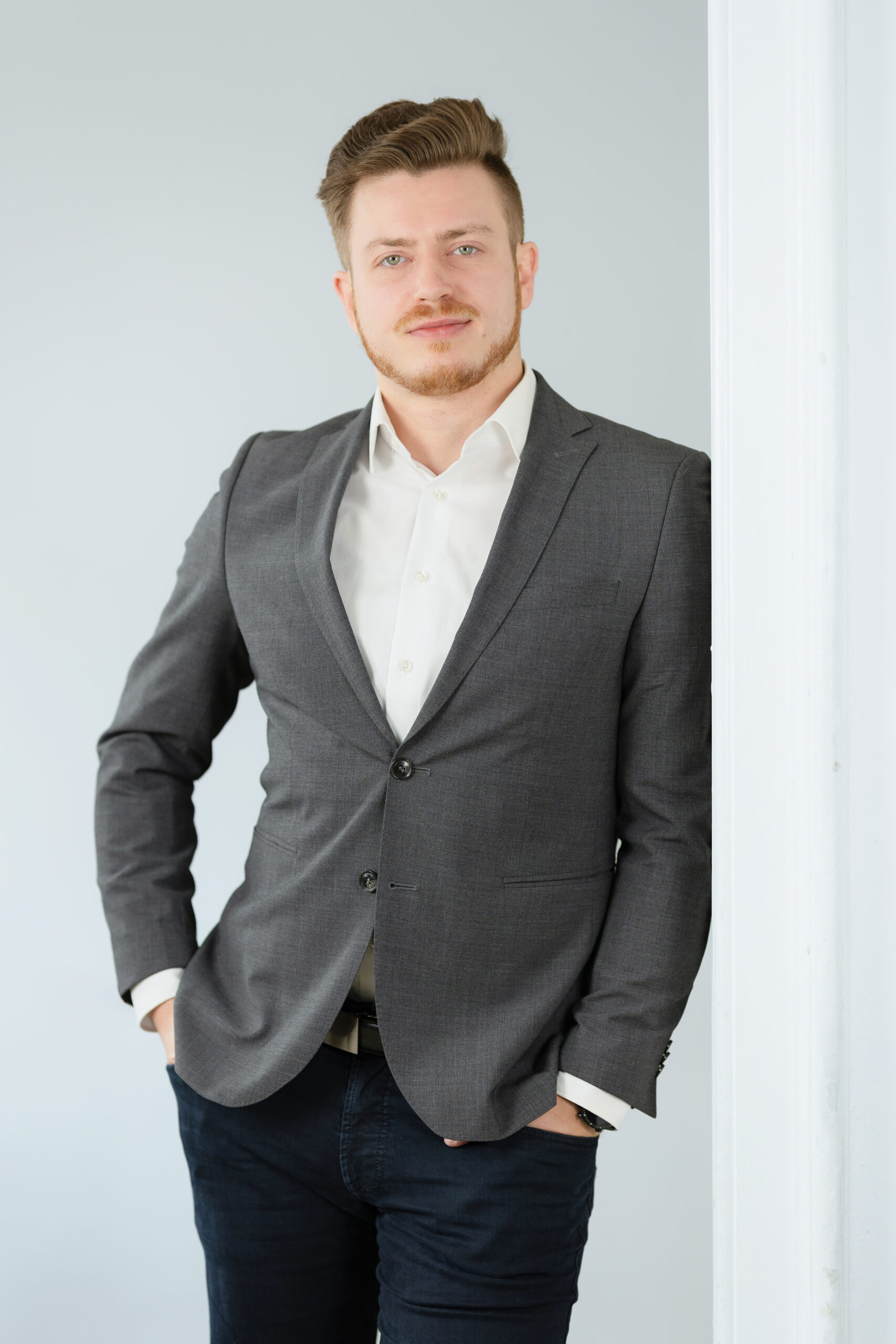 Junger Mann in weißem Hemd und grauen Anzug an die Wand gelehnt, schaut in die Kamera